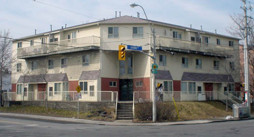 Three-storey building has angles, inset corner entrance up a flight of stairs, with cream-coloured siding, red-brick entrance walls, and balconies on the top floors