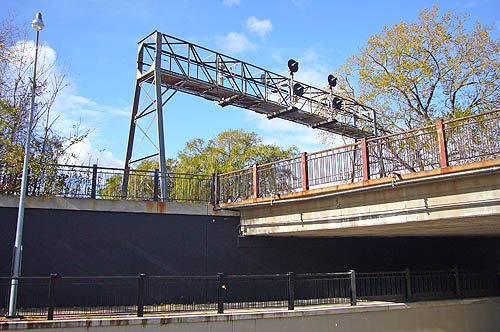 Truss holds up warning lights across a metal bridge, while underneath runs an underpass with flat black walls