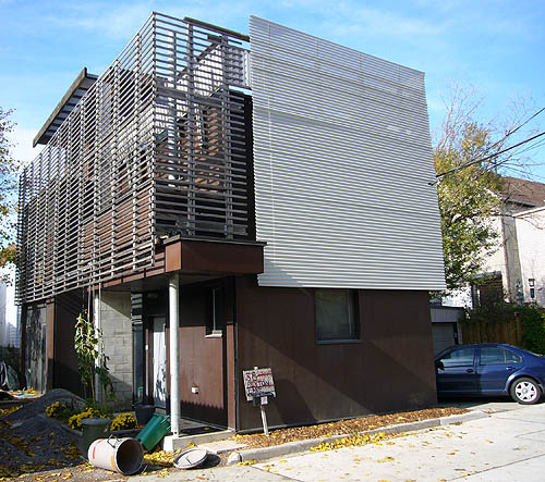 Three-floor house is covered by screens made of metal slats. One screen wraps around a two-storey side balcony