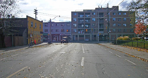 Four-lane street littered with autumn leaves terminates at a bank of three-storey buildings