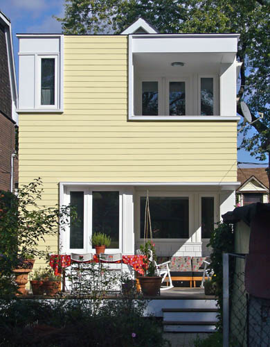 Flat wall in yellow siding has upper recessed upper deck in white trim, a lower deck in white trim, and a white-surrounded narrow window at top left