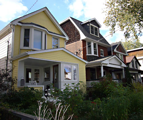 Butter-yellow house is two storeys tall, has a white-trimmed front deck with no corner column, and sits alongside brown-brick neighbours