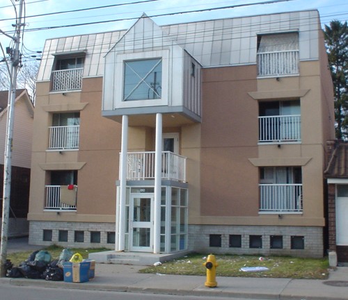 Buff-brick three-storey building has slanted aluminum top storey and a glassed-in entrance door topped by a second-floor balcony and a barn-shaped enclosed widow’s walk