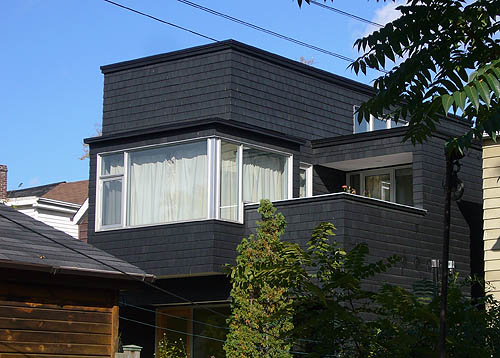 Black-shingled house has glass-windowed solarium alongside an open deck with a quarter-roof at its rear
