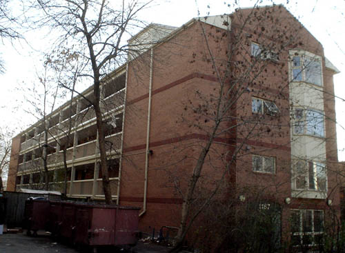 Long, narrow three-storey brown brick building has small red bands around the front and a row of balconies on all three floors on the left side