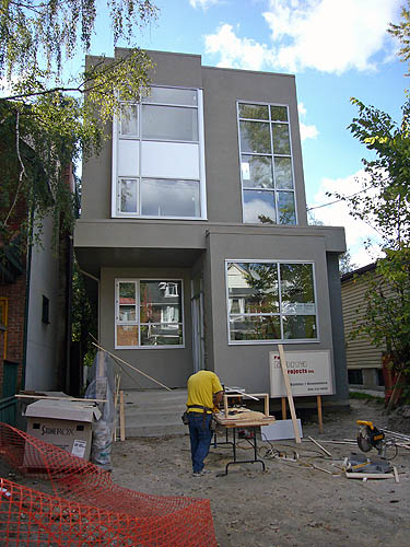 Grey-stucco house has giant windows and protruding rectangular volumes. A workman in a yellow T-shirt tends to a machine on the unfinished front yard