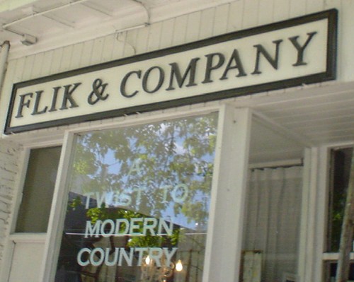 Black serif type reads FLIK & COMPANY on a white sign with black border. Letters on window read A TWIST TO MODERN COUNTRY