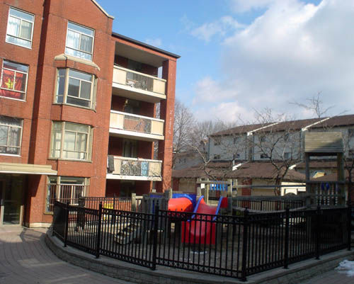 Black-fenced playgrund has red-and-blue-plastic slides and sits alongside a paved walkways leading to the bay-windowed tower of a four-storey brick building
