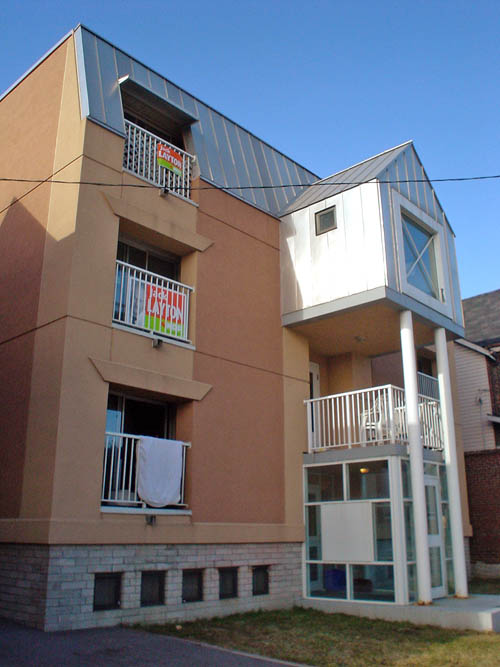 Three-quarter view sh0ows recessed balconies with white bannisters (one of which hides a Jack Layton sign) and plain brick basement with regularly-spaced square windows