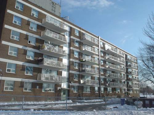 Six-storey brown-brick building has four white balconies per floor and a fence around its snowy lot