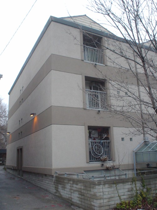 Three-quarter view of building shows bicycle and other items packed into recessed balconies, a security floodlight, and a side wall interrupted only by six punched windows at the halfway point