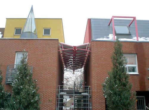 Three-storey building is divided into two brown-brick pods with a central covered entranceway. Left pod has a yellow set-back roof and a glass teepee; right pod has a slate-grey set-back roof with a red frame built around a window
