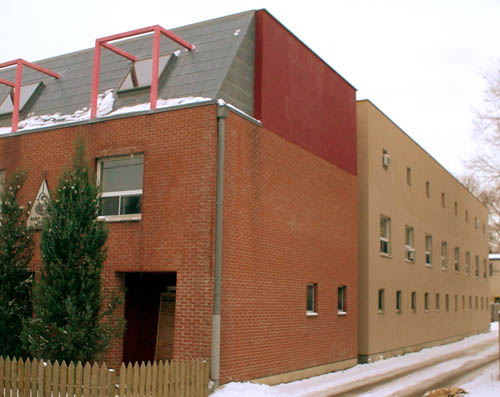 Three-quarter view shows building with, on the front section, brown-brick sides topped by red brick; a groove running the height of the building; and a buff-brick rear section with regular windows on all three floors