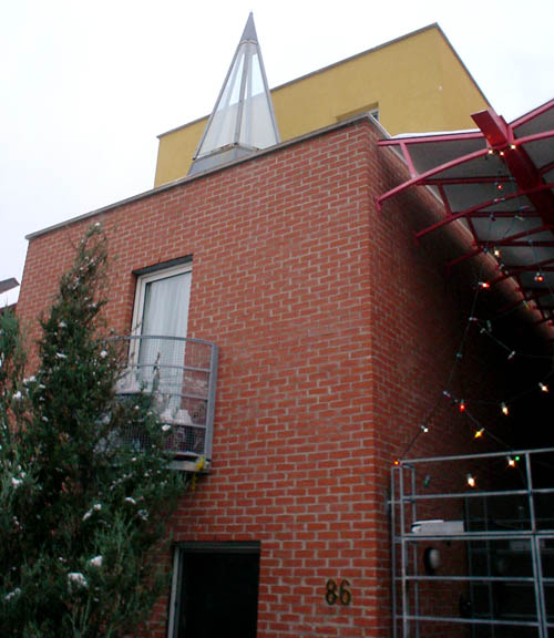 Corner view shows brown-brick building alongside a walkway with metal grated doors and glass covering. Second-floor window has semicircular Juliet balcony. Glass teepee sits in front of the set-back yellow third storey