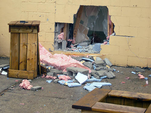 Insulation and cinder blocks sit in a pile on the ground outside a jagged hole in a yellow cinder-block wall