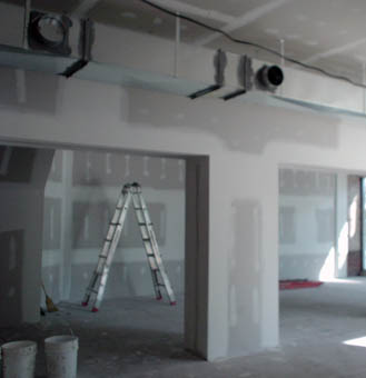 Half-painted drywall on walls and columns, pails on the floor, a disused ladder, ducts and clamps on a ceiling HVAC member