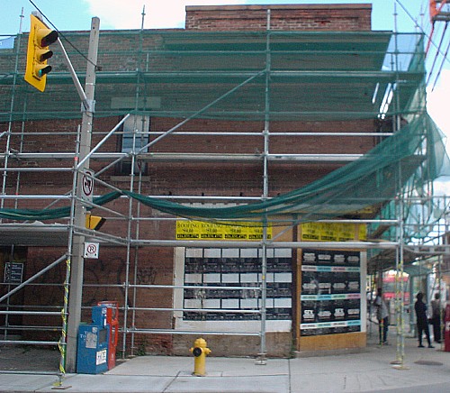 Construction scaffolding and netting surround a two-storey building whose wooden walls are covered with advertising posters