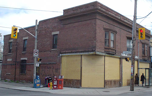 Two-storey brown brick building has plywood-covered windows on side, corner entrance, and front