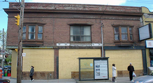 Two-storey brown brick building has two bay windows and a regular window equally spaced along on the upper floor and front windows completely covered by plywood on the ground floor