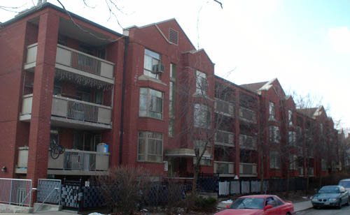 Three-storey brick building runs all the way down a street, with three taller gabled towers at regular intervals. White balconies are present on the top three floors, and the towers have bay windows